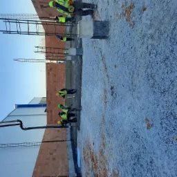 Wznoszenie muru z czerwonej cegły z wystającymi zbrojeniami, widoczni robotnicy w żółtych kamizelkach i kaskach, szary grunt i błękitne niebo w tle, wąż do pompowania betonu przy budynku.