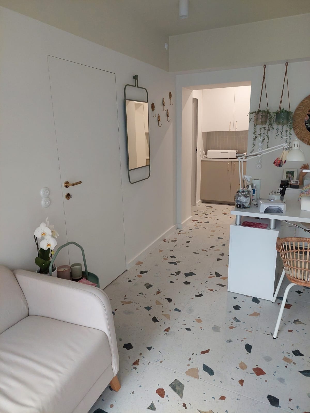 Jasne wnętrze z podłogą w stylu lastryko, białymi ścianami, ukrytymi drzwiami z złotą klamką, lustrem w metalowej ramie, fragmentem sofy i stanowiskiem do manicure z lampą oraz wiszącymi roślinami.