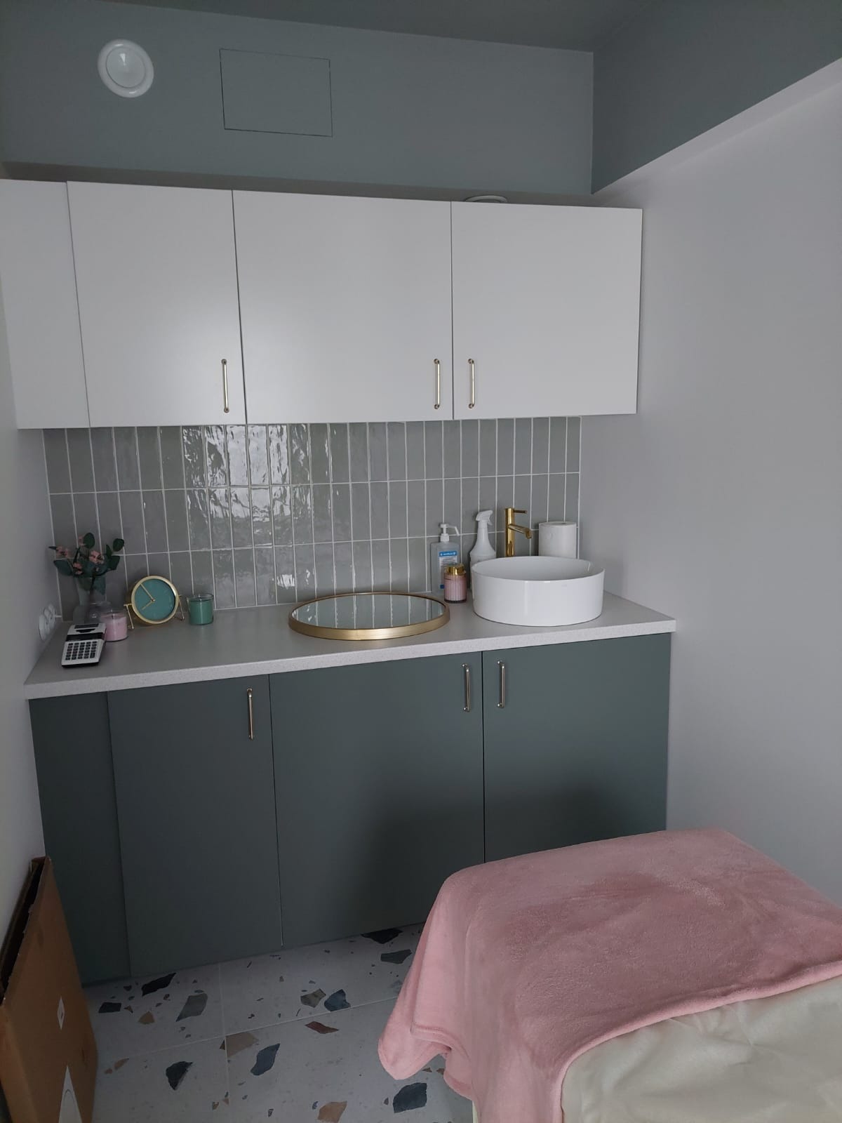 Nowoczesna łazienka z szarymi płytkami na ścianie, białymi szafkami wiszącymi i zielonymi szafkami pod umywalką nablatową, uzupełniona dekoracjami i akcesoriami w odcieniach złota i różu.