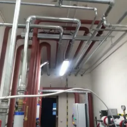 Rozbudowana sieć rur grzewczych i instalacyjnych pod sufitem w pomieszczeniu technicznym, widoczne rury stalowe w kolorze czerwonym i srebrnym, mocowania i oświetlenie ledowe.