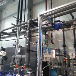 Kompleksowa instalacja przemysłowa z rurami, zaworami, czujnikami, pompami i laptopem na stole w tle, widoczne elementy instalacji wodnej i grzewczej.