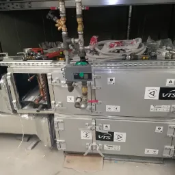 Szara centrala wentylacyjna VTS z podłączonymi rurami i zaworami, widoczne narzędzia i akcesoria na górze urządzenia.