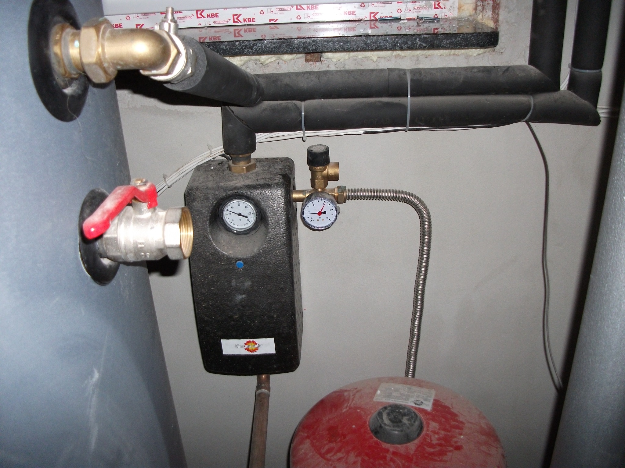 Fragment kotłowni z szarym zbiornikiem, czarnymi rurami izolowanymi termicznie, czerwonym zbiornikiem wyrównawczym i osprzętem z manometrami, widoczne ślady kurzu i zabrudzeń.