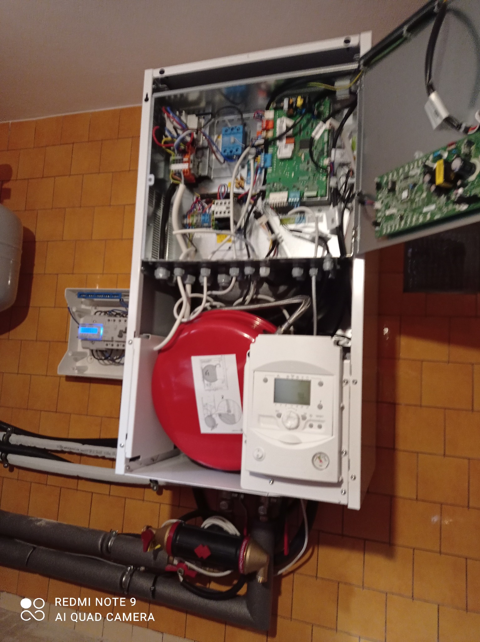 Otwarty piec gazowy wiszący na ścianie z brązowych płytek, widoczne wewnętrzne komponenty, rury instalacji grzewczej z izolacją oraz panel sterowania z wyświetlaczem.