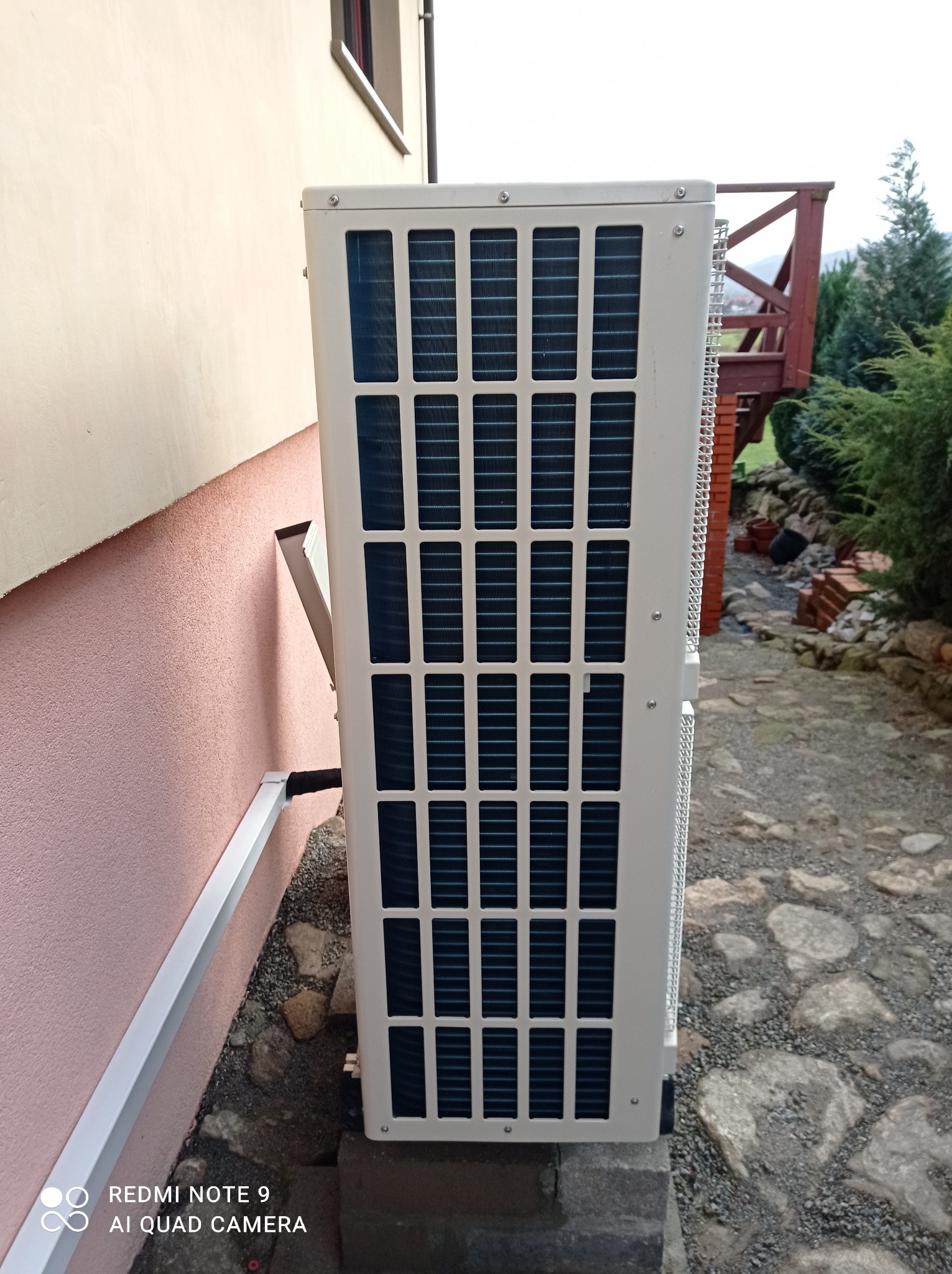 Zewnętrzna jednostka pompy ciepła zamontowana przy różowej ścianie domu, widoczny fragment kamiennej ścieżki i drewnianego tarasu w tle.