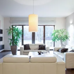 Jasny salon z białymi skórzanymi sofami, drewnianą podłogą, roślinami doniczkowymi i cylindryczną lampą wiszącą, widok na zimowy ogród przez okno.