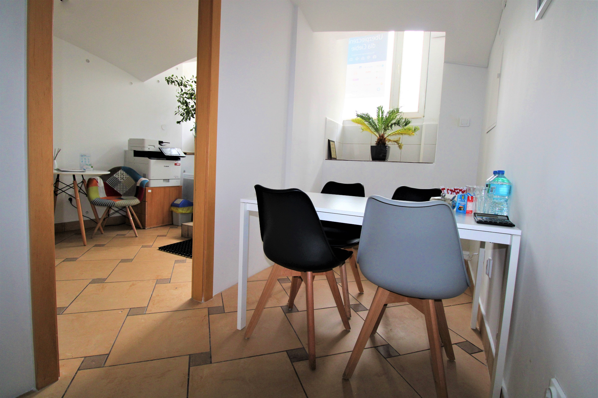 Jasne biuro z białym stołem, krzesłami w kolorze szarym i czarnym, rośliną w oknie, butelkami wody i kalkulatorem na stole, widok z korytarza.