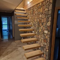 Drewniane schody wspornikowe z podświetleniem LED, zamontowane przy ścianie z kamienia dekoracyjnego. Widok na korytarz z marmurową podłogą i oknem.