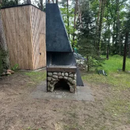 Nowoczesny grill ogrodowy z kamienia i metalu, zadaszony, na tle leśnego krajobrazu. Widoczna konstrukcja z kamienia i ciemnego metalu, obok drewniana budka.