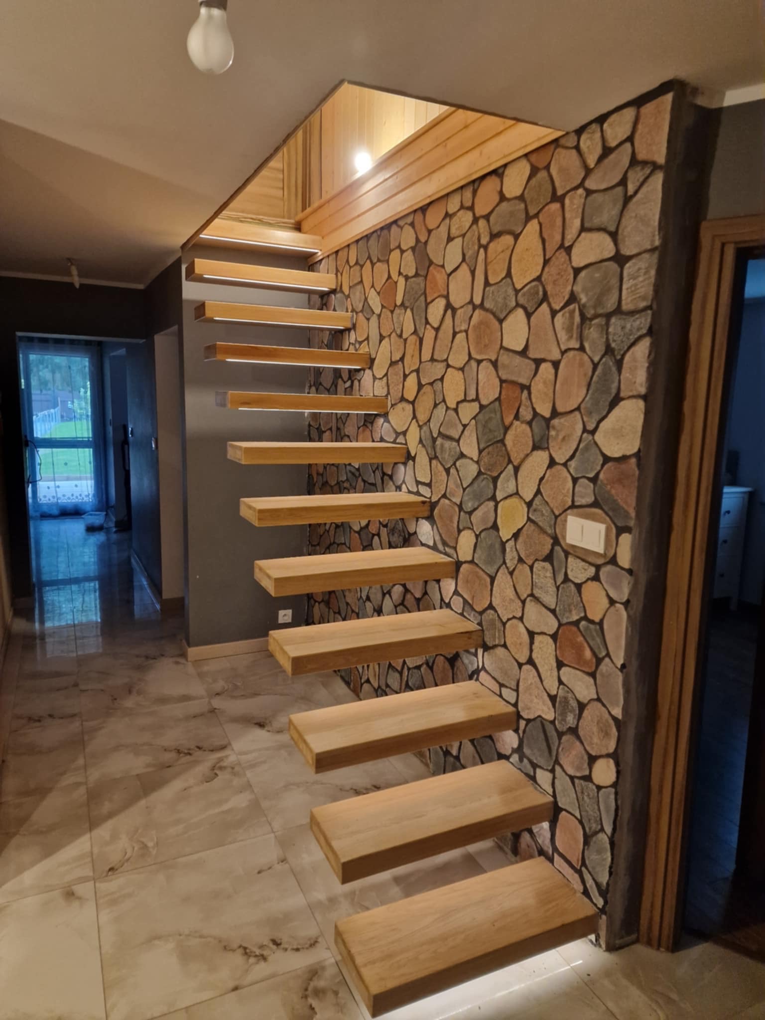 Drewniane schody wspornikowe z podświetleniem LED, zamontowane przy ścianie z kamienia dekoracyjnego. Widok na korytarz z marmurową podłogą i oknem.