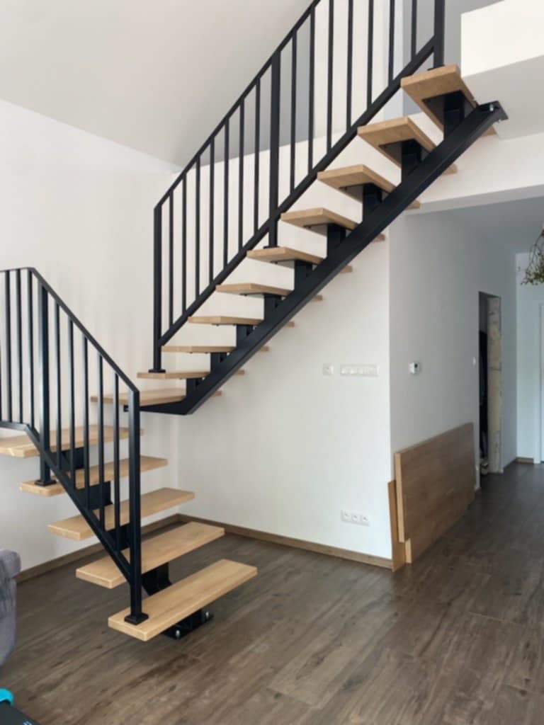 Nowoczesne schody z czarną metalową konstrukcją i jasnymi drewnianymi stopniami, widoczne w jasnym wnętrzu domu z drewnianą podłogą.