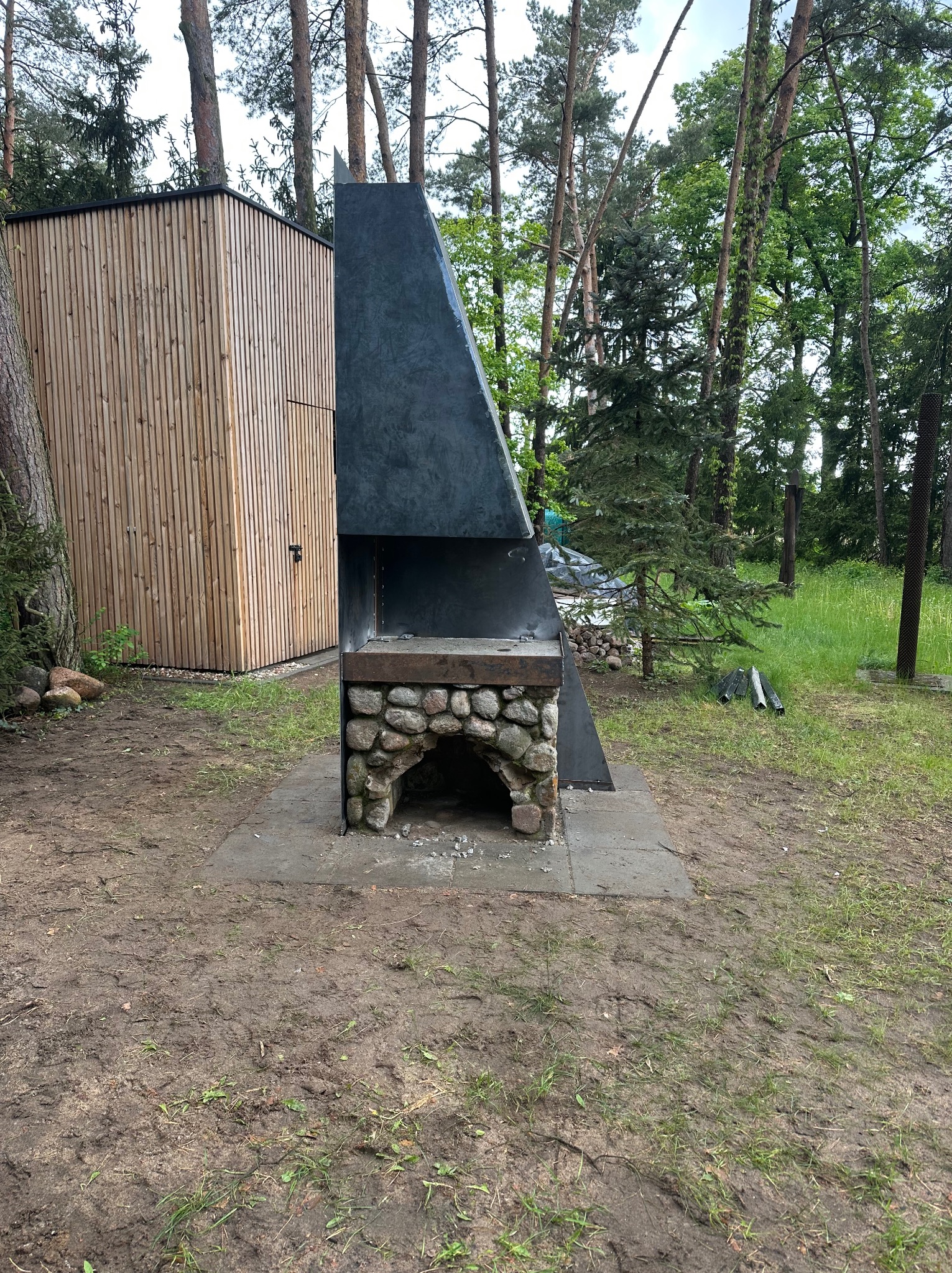 Nowoczesny grill ogrodowy z kamienia i metalu, zadaszony, na tle leśnego krajobrazu. Widoczna konstrukcja z kamienia i ciemnego metalu, obok drewniana budka.