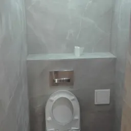 Świeżo wyremontowana toaleta podwieszana w łazience wyłożonej szarymi płytkami imitującymi marmur, z ukrytym spłukiwaniem i rolką papieru toaletowego na półce.