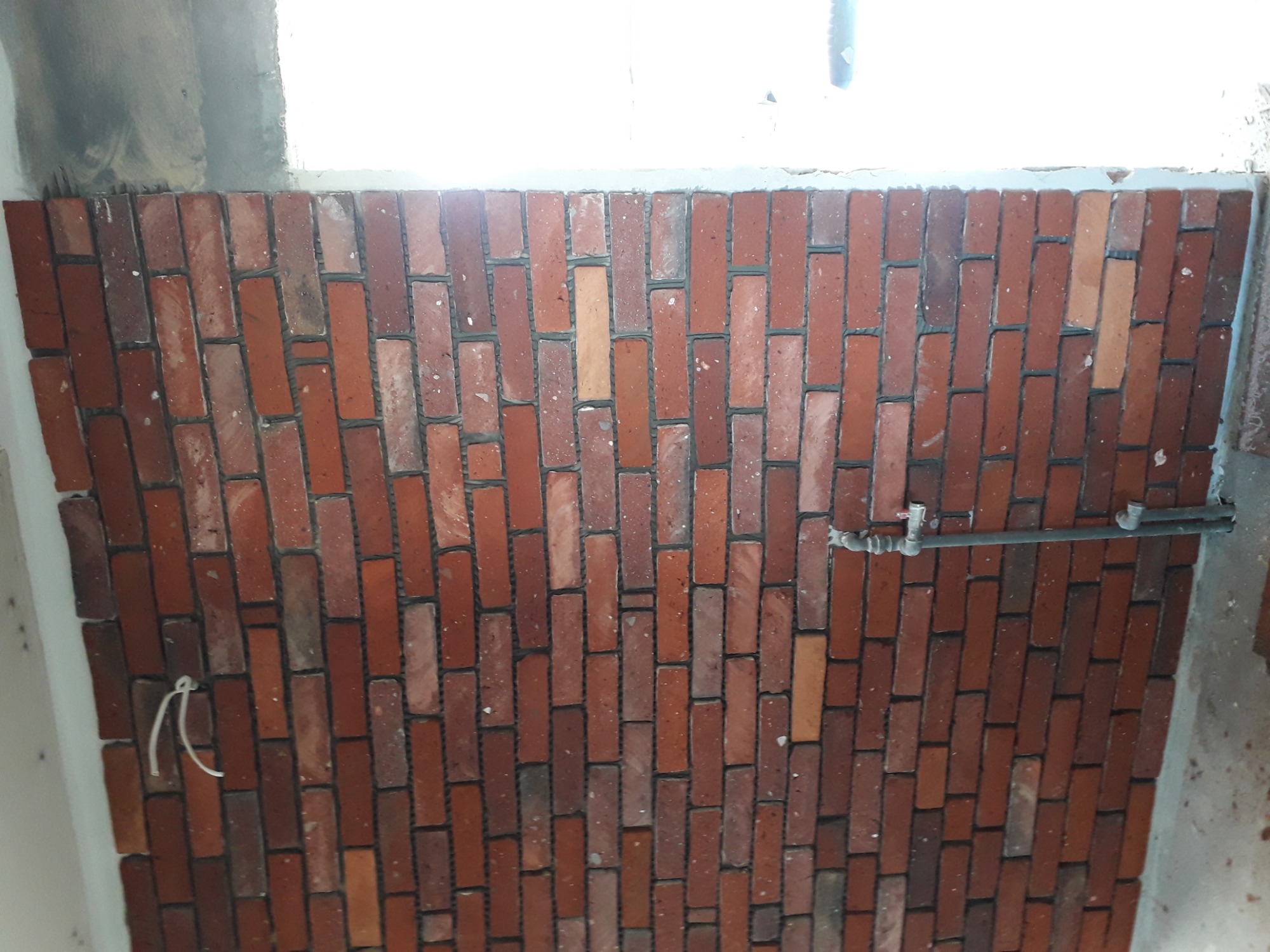Ściana wyłożona czerwoną cegłą klinkierową z widoczną instalacją hydrauliczną w tle, wykonana w trakcie remontu.