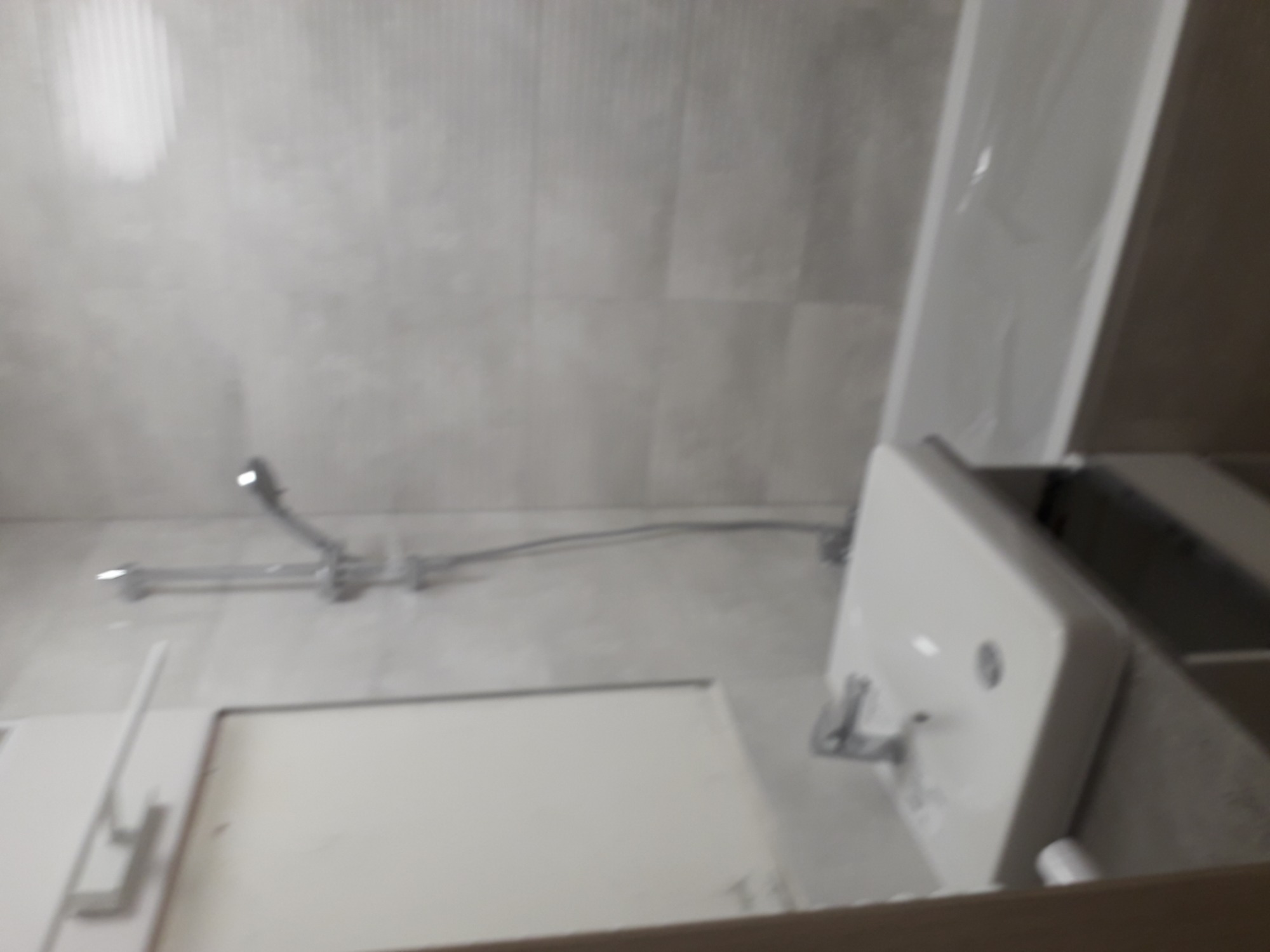 Szare płytki na ścianie łazienki, bateria prysznicowa z wężem, biała szafka z umywalką w trakcie montażu, częściowo zakryta folią ochronną.