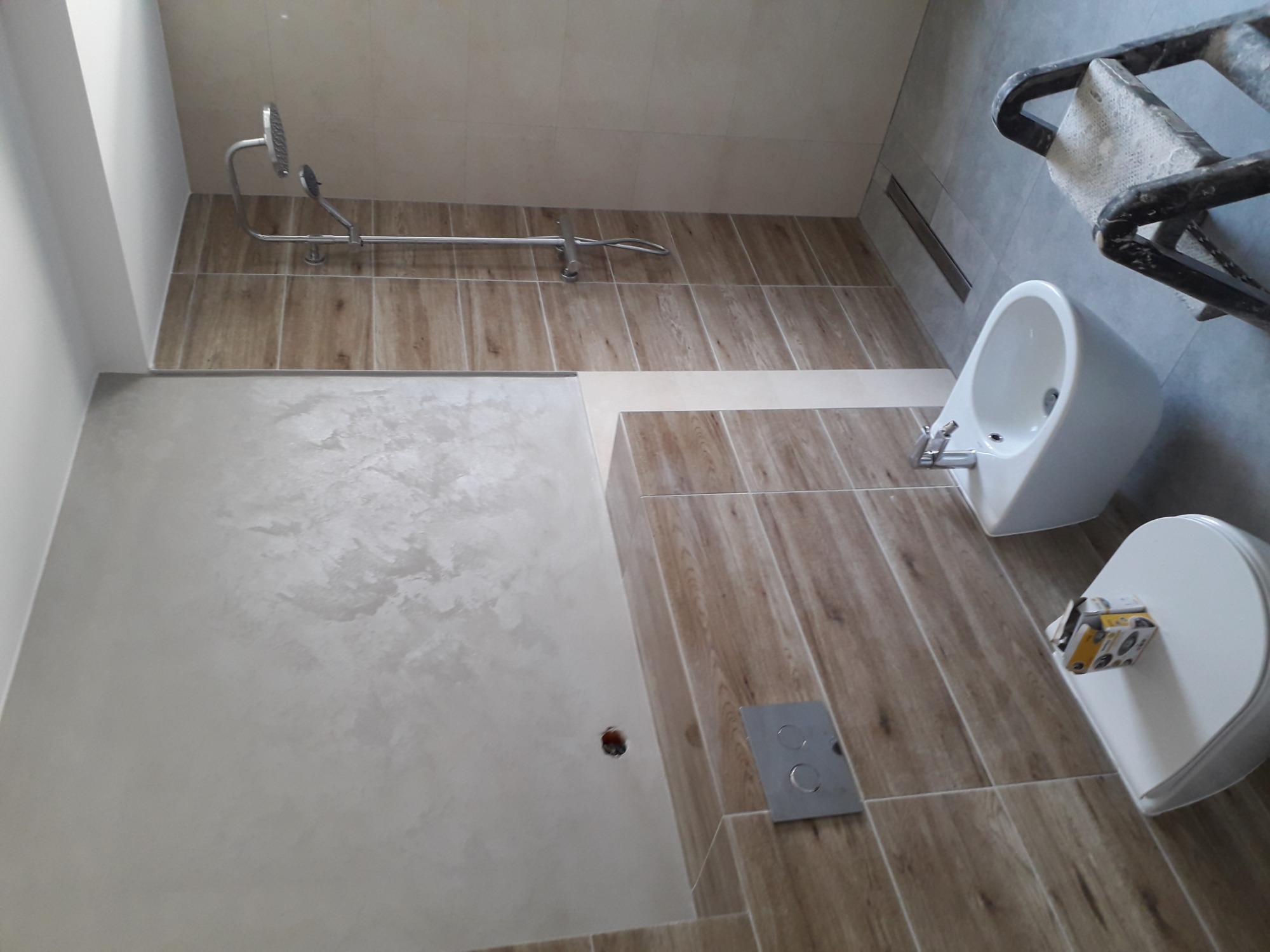 Wykończona łazienka z drewnopodobnymi płytkami, bidetem, toaletą i prysznicem; widoczny fragment drabiny malarskiej.