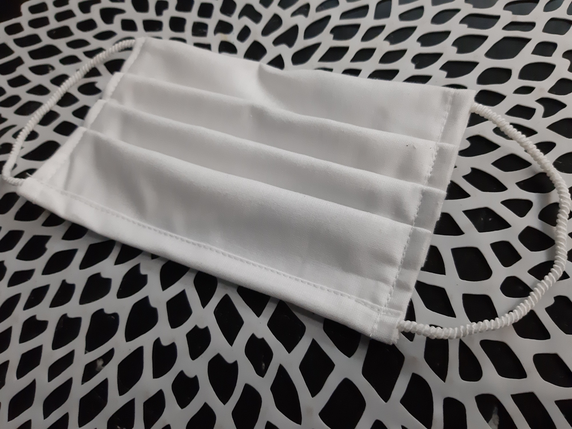 Biała, plisowana maska ochronna z elastycznymi gumkami, leżąca na ażurowej, białej powierzchni.