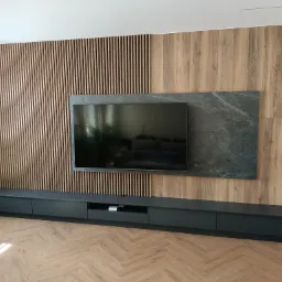 Nowoczesna ściana TV z drewnianymi lamelami i panelem z efektem kamienia. Długa, czarna szafka RTV na parkiecie w jodełkę. Minimalistyczny design wnętrza.