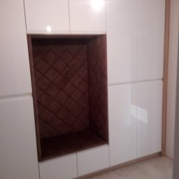 Meblomar-mikstat - Biała szafa wnękowa z brązową, pikowaną wnęką do siedzenia, obramowana drewnianą listwą. Błyszczące fronty szafek, jasna podłoga.
