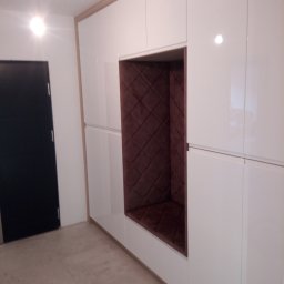 Meblomar-mikstat - Biała szafa wnękowa z brązowym, tapicerowanym siedziskiem wnękowym. Minimalistyczny design, gładkie fronty, widoczna czarna krawędź drzwi po lewej.