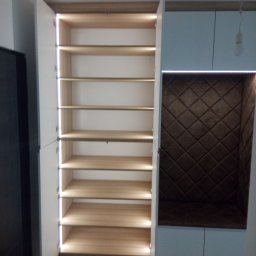 Meblomar-mikstat - Biała szafa wnękowa z otwartymi drzwiami, podświetlane półki z drewna, obok siedzisko z brązowym pikowanym oparciem i podświetleniem LED.