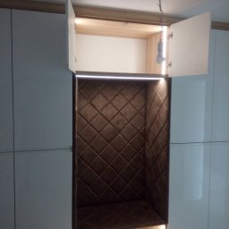 Meblomar-mikstat - Biała szafa na wymiar z otwartą wnęką wyłożoną brązową, pikowaną tkaniną. W dolnej części wnęki widoczne dwie wysuwane szuflady z oświetleniem LED.