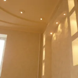 Wykończony pokój z dekoracyjną ścianą z wnękami oświetlonymi punktowo, wzorzysta struktura ściany, podwieszany sufit z oświetleniem halogenowym, okno z widocznymi przedmiotami na parapecie.