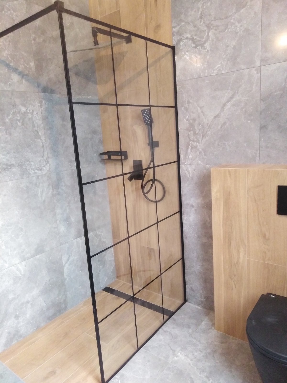 Nowoczesna kabina prysznicowa walk-in z czarnymi profilami, deszczownicą i baterią w kolorze czarnym matowym, na tle płytek imitujących drewno i marmur.