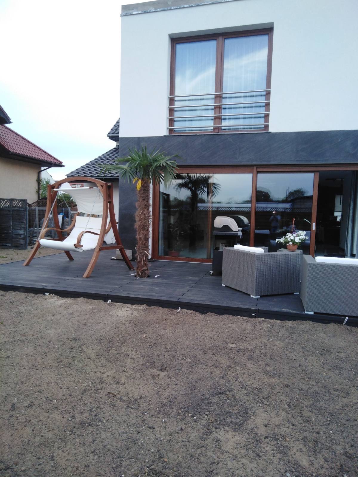 Nowoczesny taras z ciemnej deski kompozytowej, huśtawką ogrodową typu kokon, szarymi meblami wypoczynkowymi i małą palmą, przylegający do domu z dużym oknem przesuwnym.