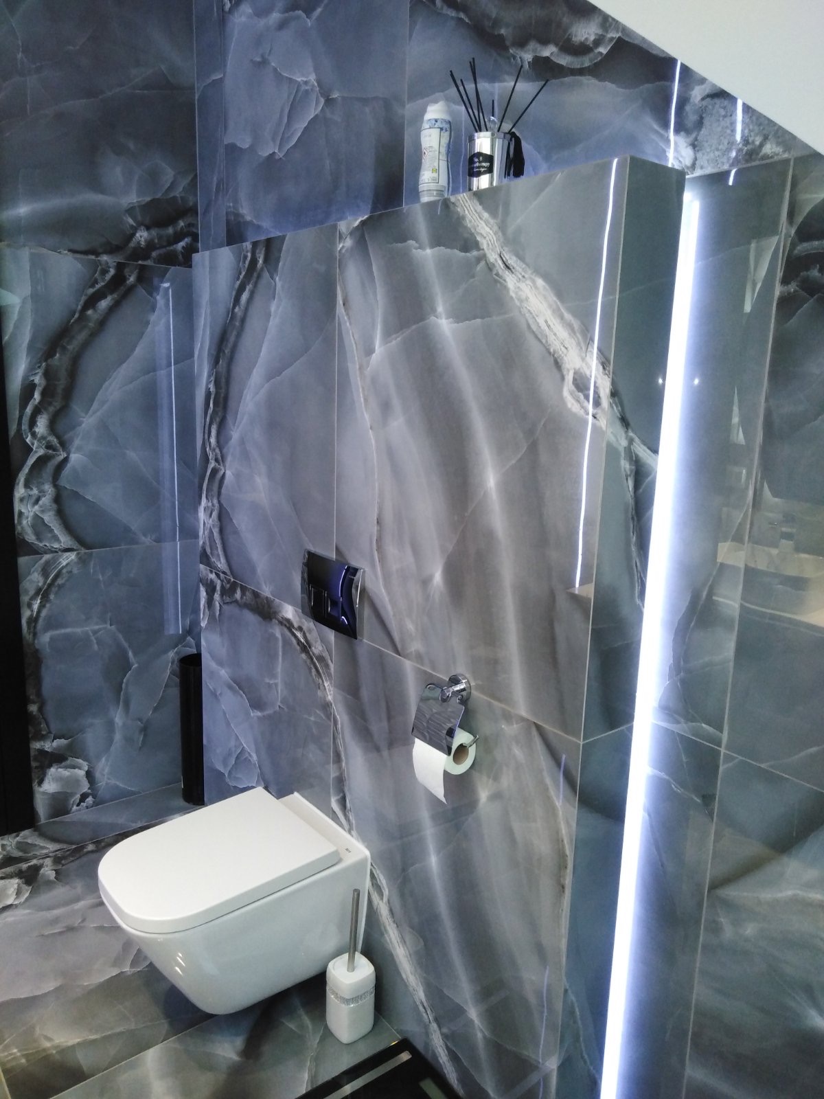 Nowoczesna łazienka z szarymi płytkami imitującymi marmur, podwieszana toaleta i oświetlenie LED w pionowych pasach.