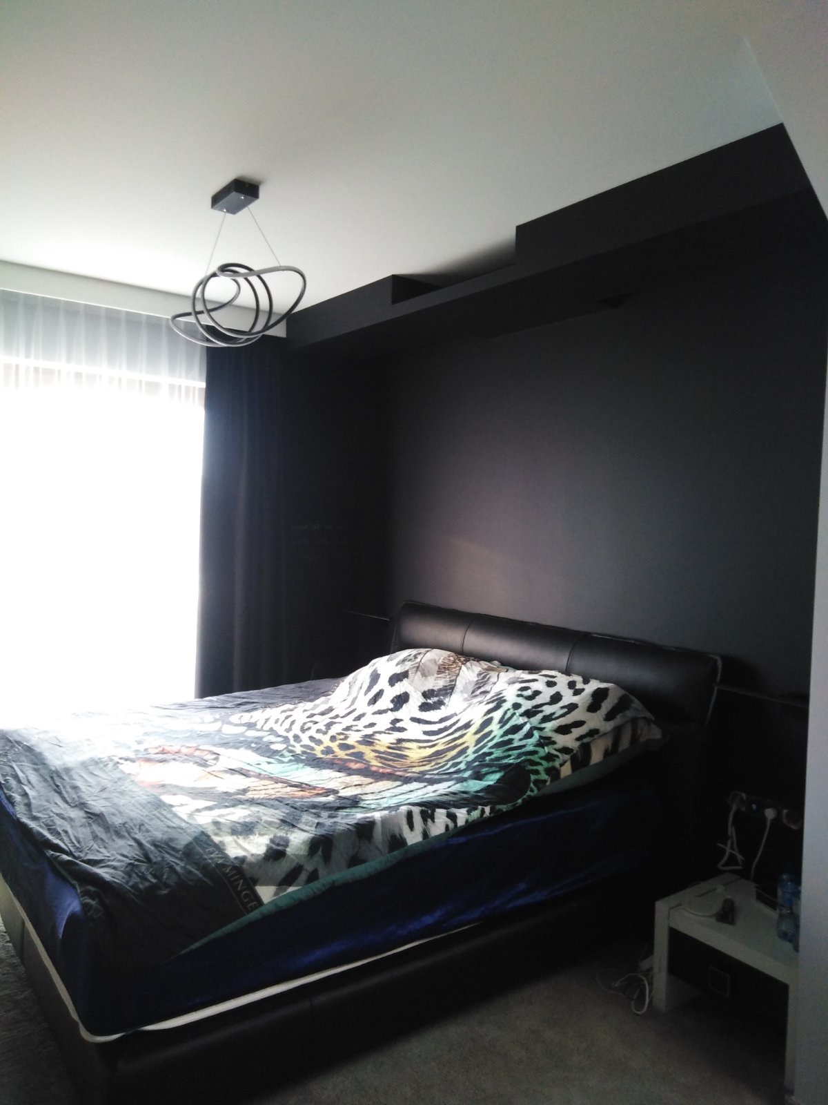 Czarny pokój sypialny z łóżkiem z czarną ramą i wzorzystą narzutą, nowoczesna lampa wisząca i czarne ściany z wnęką.