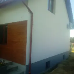 Dwukondygnacyjny dom z ocieploną elewacją, częściowo wykończoną drewnem, z widoczną rynną i klimatyzatorem na ścianie.