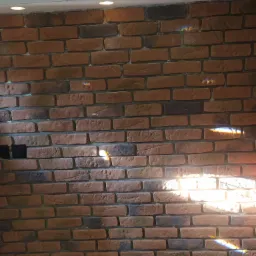 Dekoracyjna ściana z cegły klinkierowej w odcieniach brązu i szarości, oświetlona punktowymi lampami sufitowymi i promieniami słonecznymi tworzącymi tęczowe refleksy.