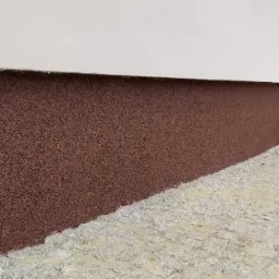 Dolna część ściany budynku pokryta brązowym tynkiem mozaikowym, kontrastująca z białą górną częścią ściany i szarą kostką brukową wzdłuż fundamentów.