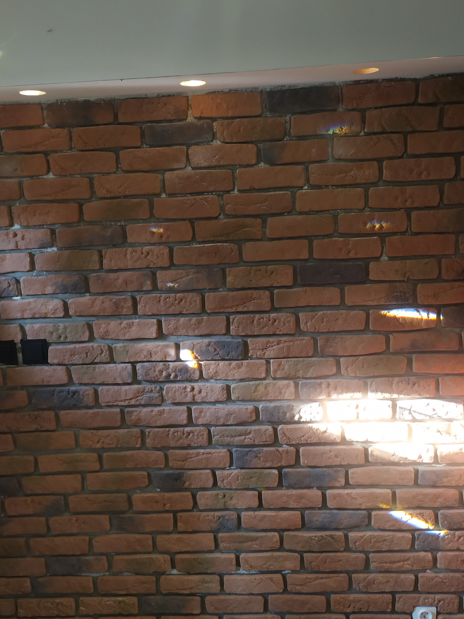 Dekoracyjna ściana z cegły klinkierowej w odcieniach brązu i szarości, oświetlona punktowymi lampami sufitowymi i promieniami słonecznymi tworzącymi tęczowe refleksy.