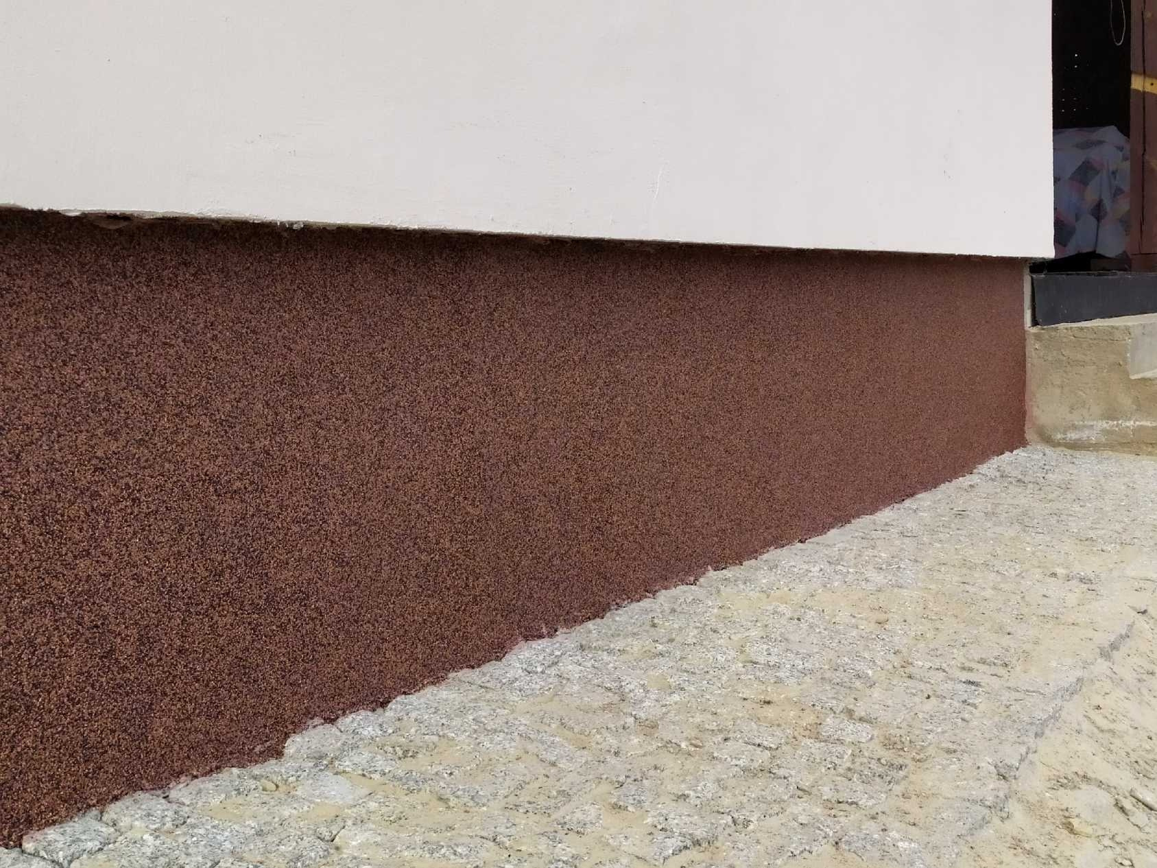 Dolna część ściany budynku pokryta brązowym tynkiem mozaikowym, kontrastująca z białą górną częścią ściany i szarą kostką brukową wzdłuż fundamentów.