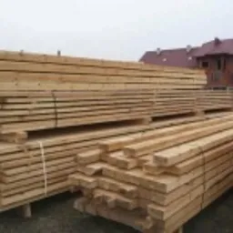 Stosy świeżo przetartego drewna na paletach, widoczne słoje i jasny kolor, w tle budynki z czerwonymi dachami.