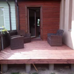 Drewniany taras z brązowymi krzesłami rattanowymi, częściowo zadaszony, widok na otwarte drzwi domu, narzędzia i materiały budowlane na trawie poniżej.