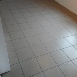 Ułożone płytki podłogowe w pustym, jasnym pomieszczeniu z białymi ścianami, widoczne gniazdko elektryczne. Szare fugi podkreślają kwadratowy wzór płytek.