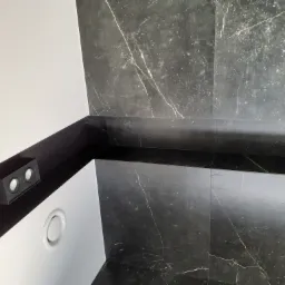 Ciemne, marmurowe płytki na ścianie i podłodze w łazience, widoczny fragment czarnej półki z oświetleniem punktowym i biały nawiewnik sufitowy.