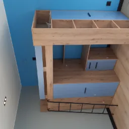 Drewniane łóżko piętrowe z półkami i drabinką, w pokoju z błękitnym sufitem i szarą ścianą, widok z dołu.