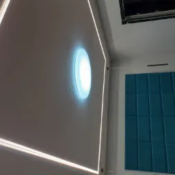 Nowoczesny sufit z oświetleniem LED po obwodzie i okrągłą lampą w centralnej części, obok dekoracyjna ściana z miękkimi panelami w kolorze turkusowym.