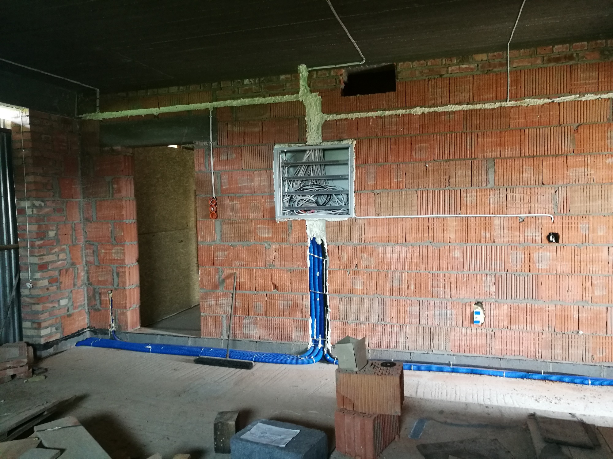 Rozdzielnia elektryczna z poprowadzonymi przewodami w nieotynkowanym pomieszczeniu z cegły, widoczne niebieskie rury instalacyjne w podłodze.
