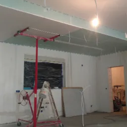 Montaż płyty gipsowo-kartonowej na suficie z użyciem podnośnika, widoczne przygotowane ściany i narzędzia w surowym wnętrzu.
