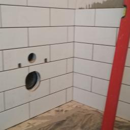 Reformas Generales - Fragment ściany wyłożonej białymi, prostokątnymi płytkami z ciemną fugą, z wyciętymi otworami na instalacje, widoczny czerwony poziomnica.