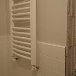 Reformas Generales - Biały grzejnik drabinkowy na ścianie wyłożonej białymi, prostokątnymi płytkami w trakcie remontu łazienki, z widocznym fragmentem surowej ściany i osobą w tle trzymającą narzędzia.