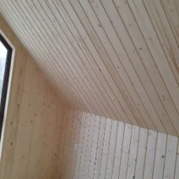 Wykończone poddasze z boazerią drewnianą na ścianach i skośnym suficie, widoczna instalacja elektryczna na suficie i gniazdko na ścianie, podłoga wyłożona panelami.