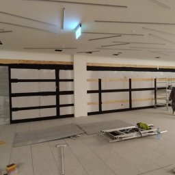 Reformas Generales - Wnętrze w trakcie montażu ścianek działowych z płyt gipsowo-kartonowych, widoczne stelaże, płyty, narzędzia i drabina, sufit z nietypowym oświetleniem.