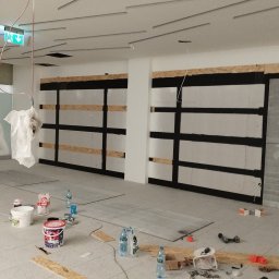 Reformas Generales - Montaż płyt gipsowo-kartonowych na ścianie, widoczne profile, płyty i materiały budowlane, w tle sufit z elementami konstrukcyjnymi, na podłodze rozłożone narzędzia i butelki z wodą.