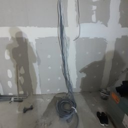 Reformas Generales - Wnętrze w trakcie prac wykończeniowych: ściany z płyt kartonowo-gipsowych, widoczne przewody elektryczne zwisające z metalowej szyny, narzędzia i buty robocze na podłodze.